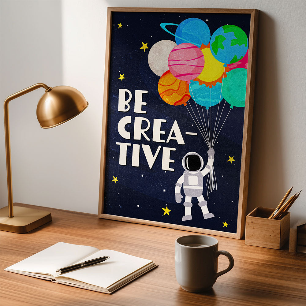A close-up shot of a desk with a poster in the center. The poster has a wooden frame and reads "BE CREATIVE." It shows a cartoon astronaut holding a bunch of balloons shaped like planets, set against a dark, starry background.