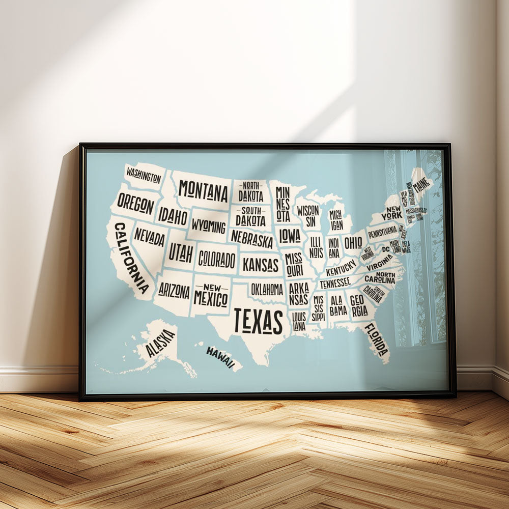 Framed map of the United States with state names on a light blue background, leaning against a white wall.