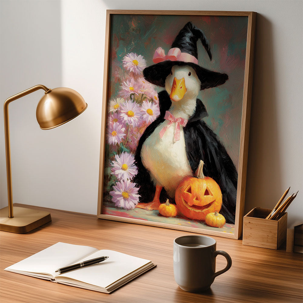 A framed artwork sits on a desk, depicting a duck in a witch costume and pumpkins.