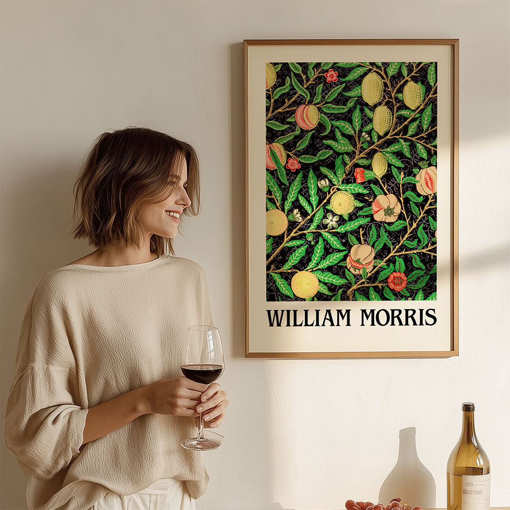 Woman holding a glass of wine, standing next to a framed William Morris artwork on a wall.